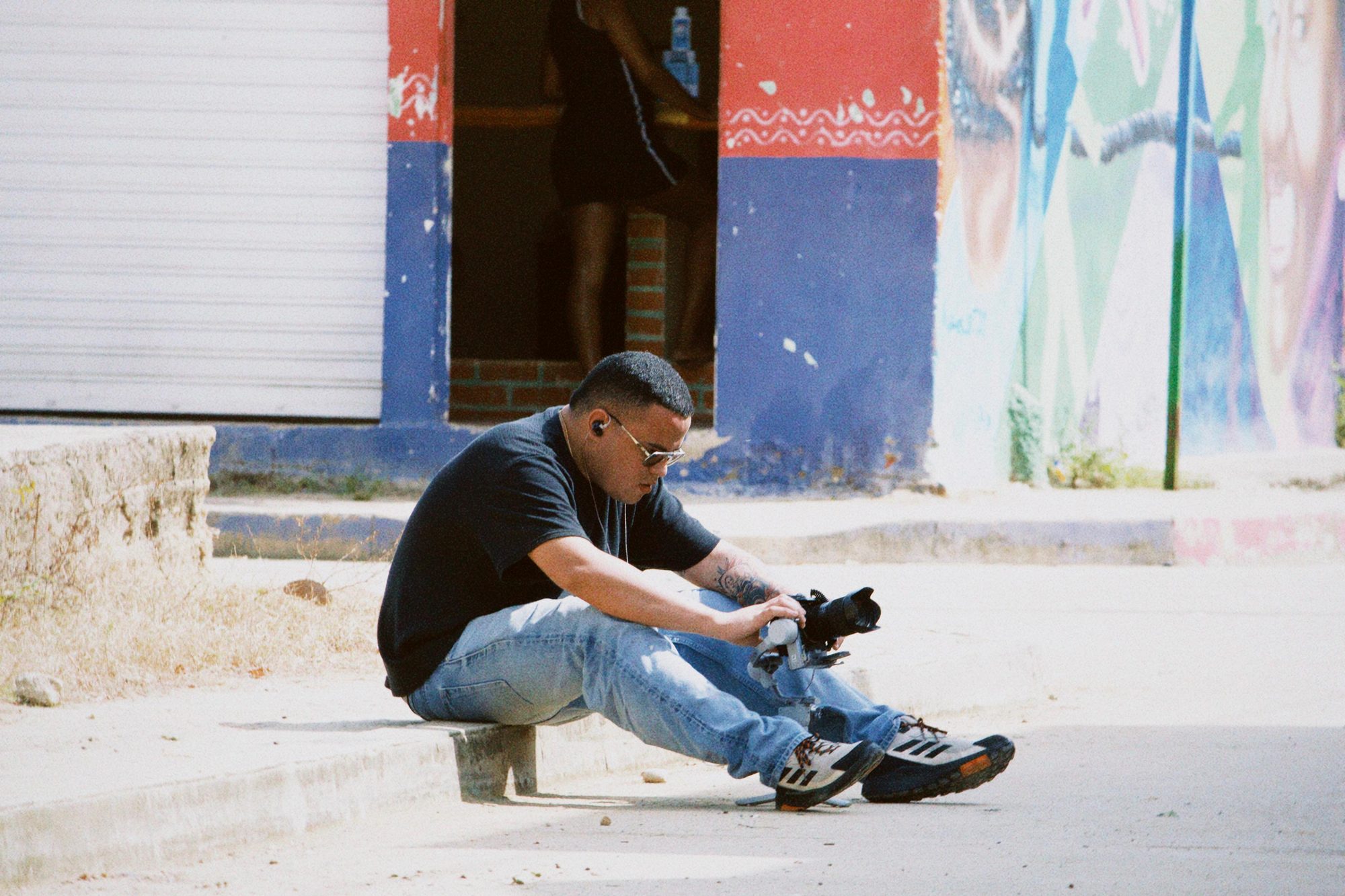 Camilo ajustando un estabilizador de cámara sentado en una acera junto a un mural