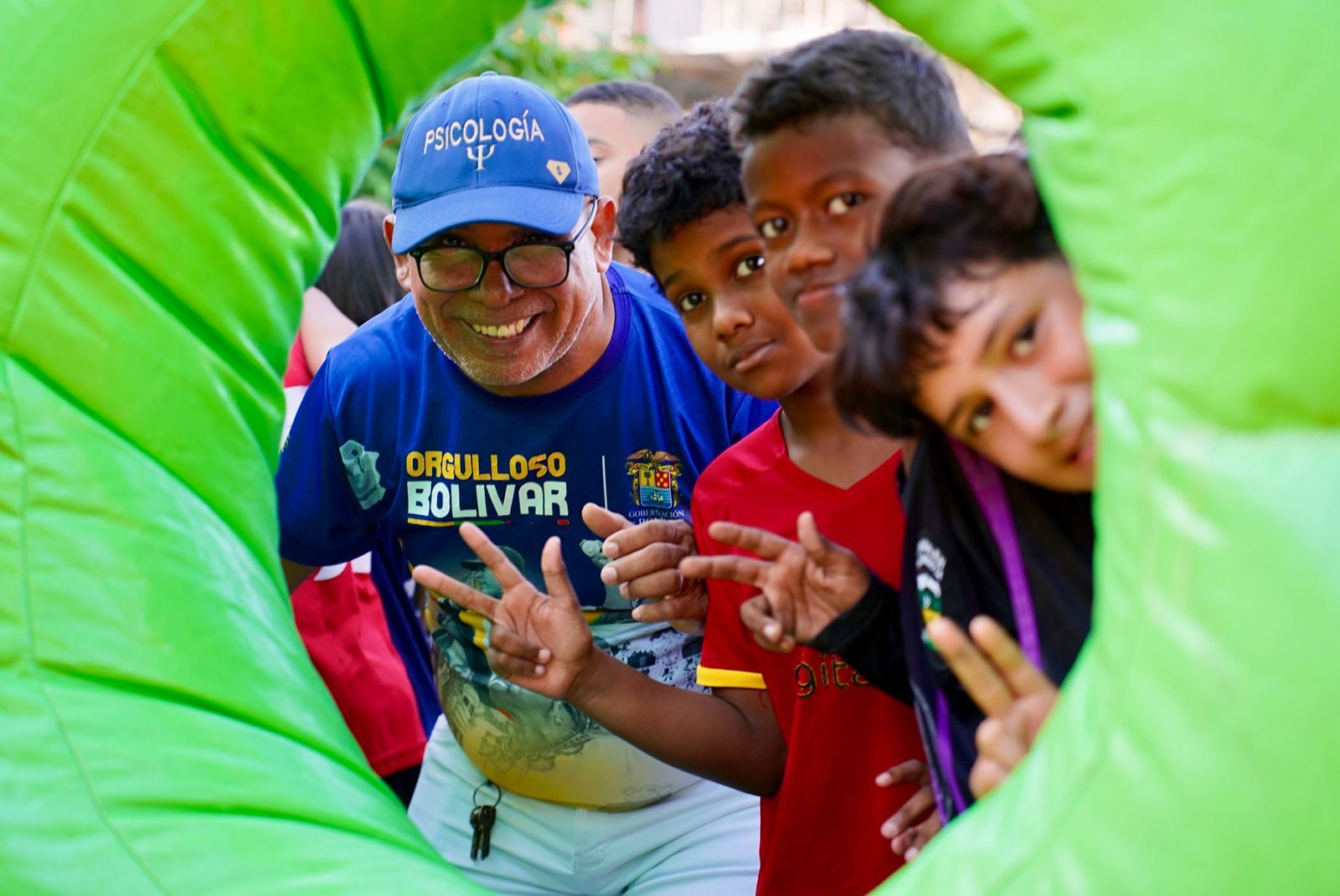 Hombre con gorra de Psicología y niños haciendo la señal de paz dentro de un túnel inflable verde