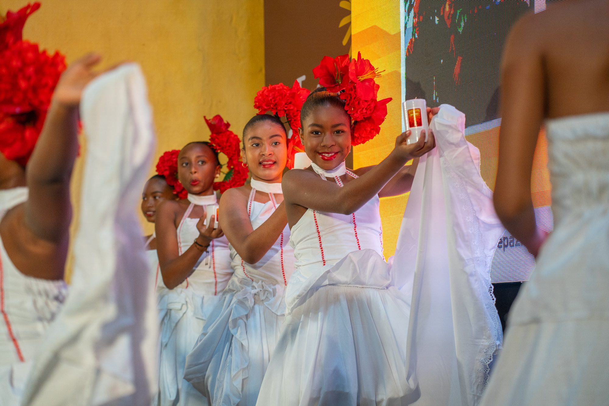 Niñas con vestidos blancos y tocados de flores rojas bailando con velas en un evento del Tour Filantrópico
